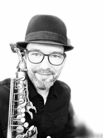 Ryan Grist, accomplished woodwind multi-instrumentalist specializing in flute, clarinet, and saxophone from Orangeville, ON, in a monochrome image. Dressed in a stylish black hat and button-up shirt, sporting glasses and a signature smile, while holding his Yamaha 82z Alto Saxophone.